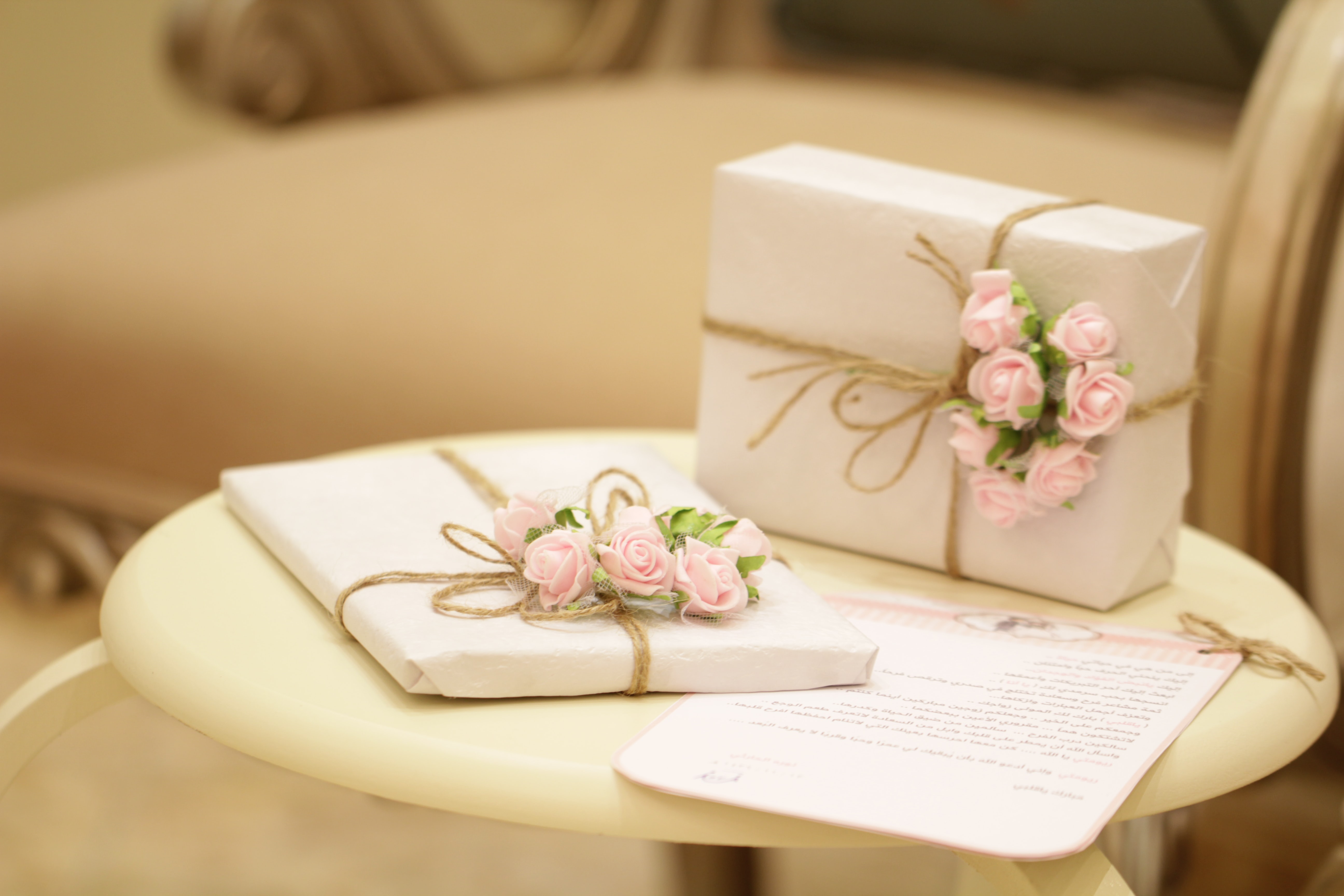 Promising Signs: Wedding Guests are in for an expensive year attending weddings. Image shows 2 gifts on a white table. Each gift is wrapped in white paper and tied with string and pink roses.